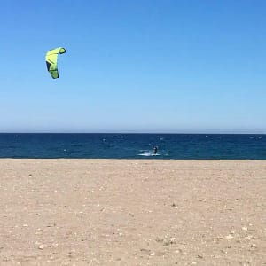 A beach in Mojácar with a kitesurfer with a green kite. The sky is blue, the sea is a deeper blue and the sand in the foreground has some small pebbles, but is mainly sand.