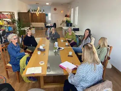 A group of eight women sit around a large wooden table in a bright, modern coworking space. The room features white walls, wooden floors, bookshelves filled with colorful books, and lush green plants. The women appear engaged in conversation, with coffee cups, notebooks, and folders on the table, suggesting a collaborative meeting or workshop. The atmosphere is warm, inclusive, and thoughtfully designed.